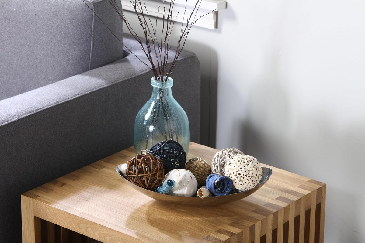 Hammered stainless steel oval bowl styled on a wooden side table holding decorative orbs beside a glass vase
