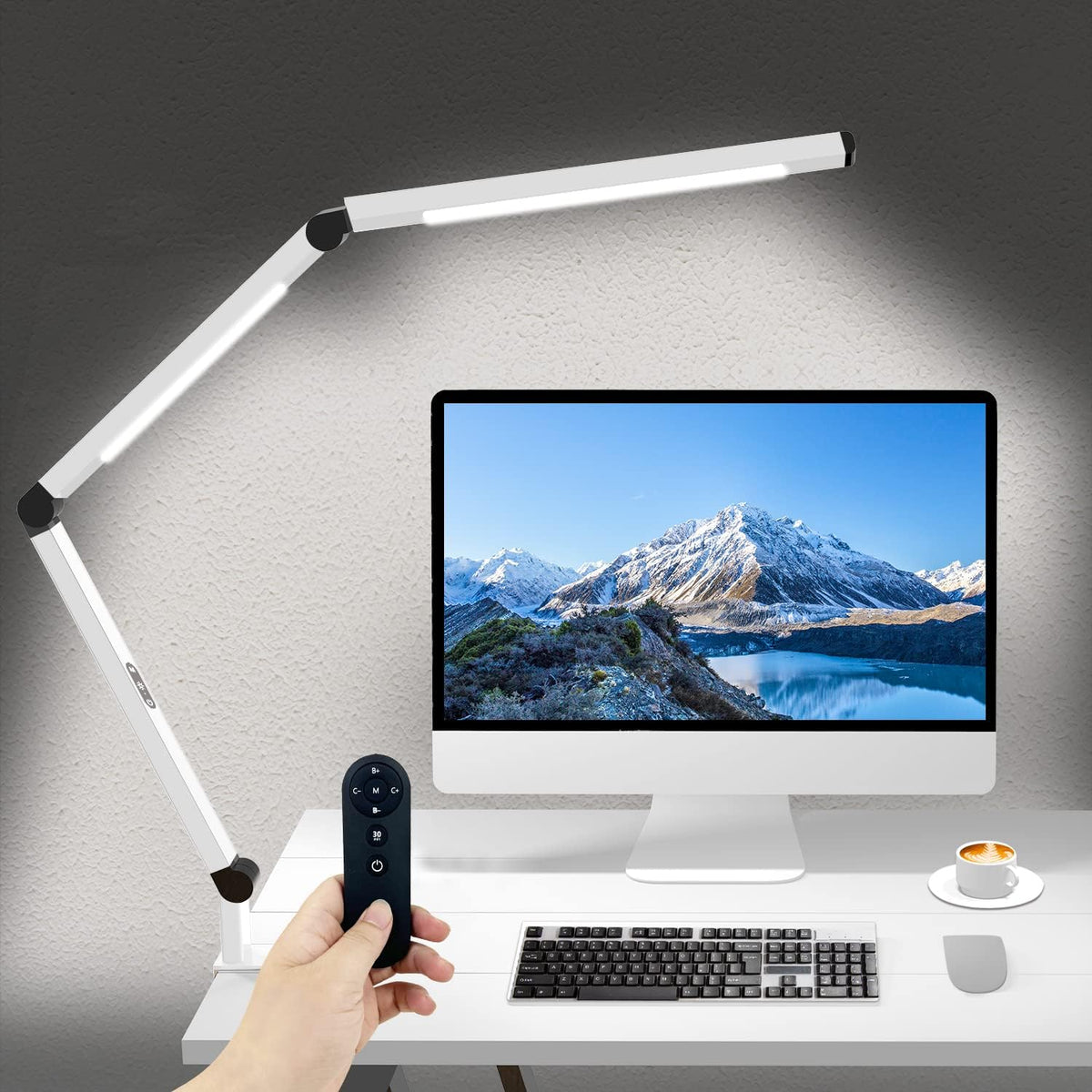 White dual-arm clamp lamp lit above a desktop monitor; hand holds the black remote in foreground.