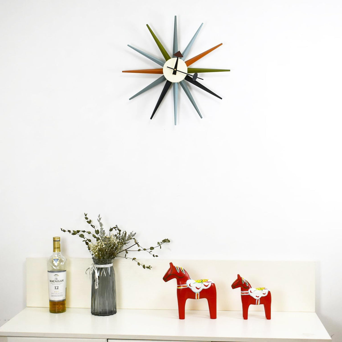 Clock mounted high on a white wall above a shelf with vase, bottle and two red horse figurines.