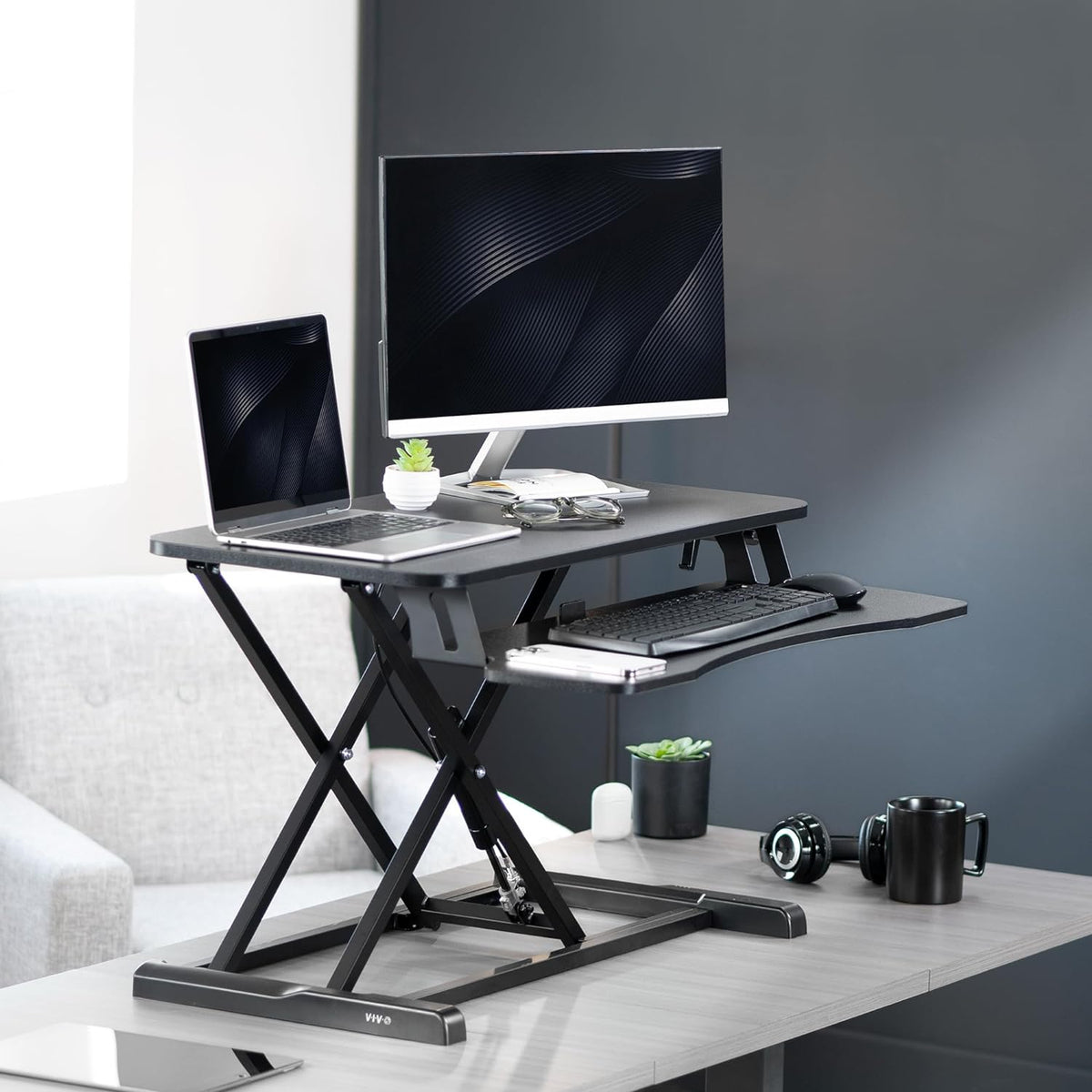 Raised black standing desk converter on a desk with a monitor, laptop, keyboard and small plants in a living room setting.