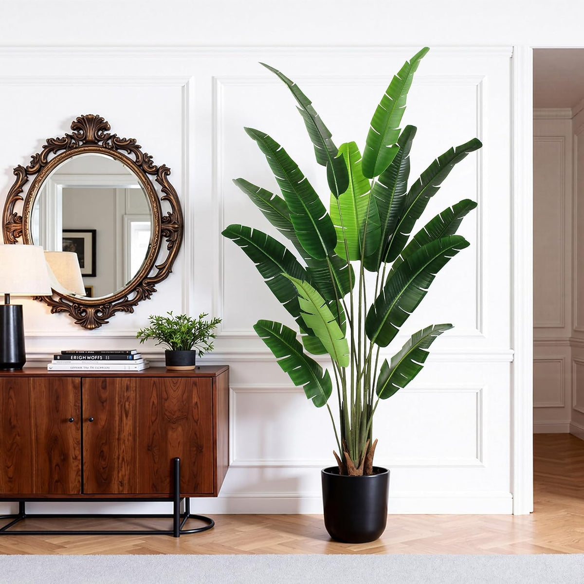 Bird of paradise tree with tall silk leaves in a black round pot beside a wooden console and mirror in a living room