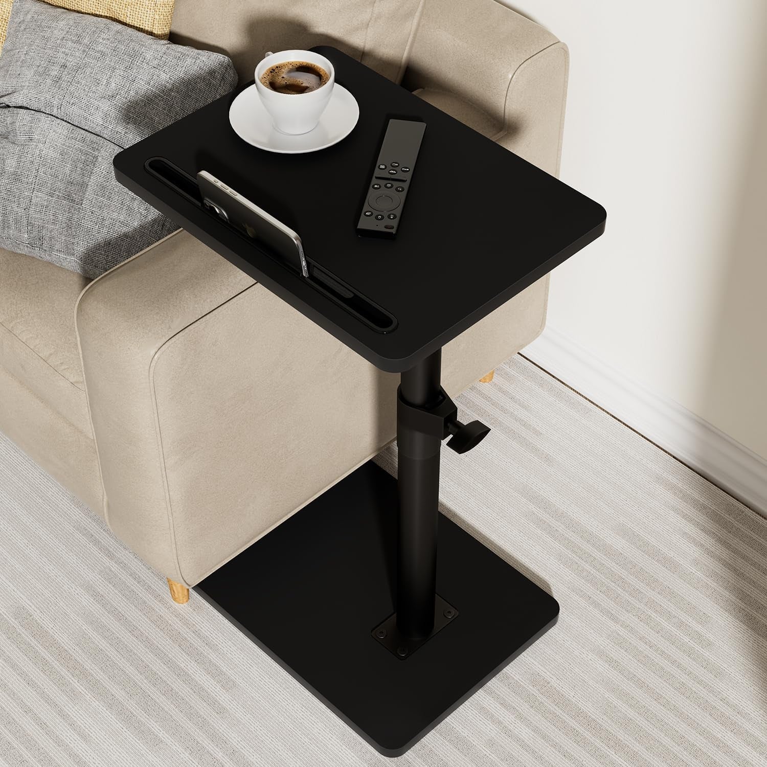 Black C‑shaped tray table slid under a sofa arm, holding a cup, remote, and phone slot.