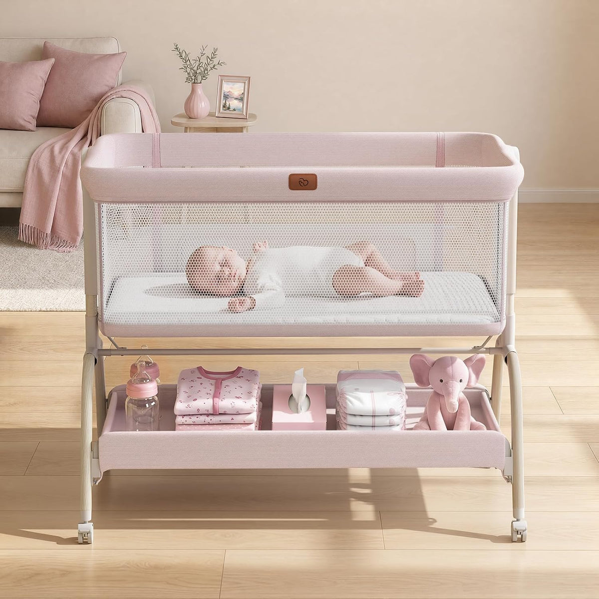 Pink bedside bassinet with mesh sides, baby inside and lower storage shelf holding baby items.