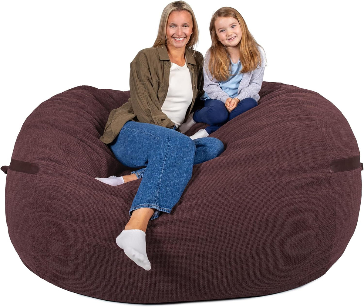 A woman and young girl sitting together on a large round brown bean bag chair on a white background.