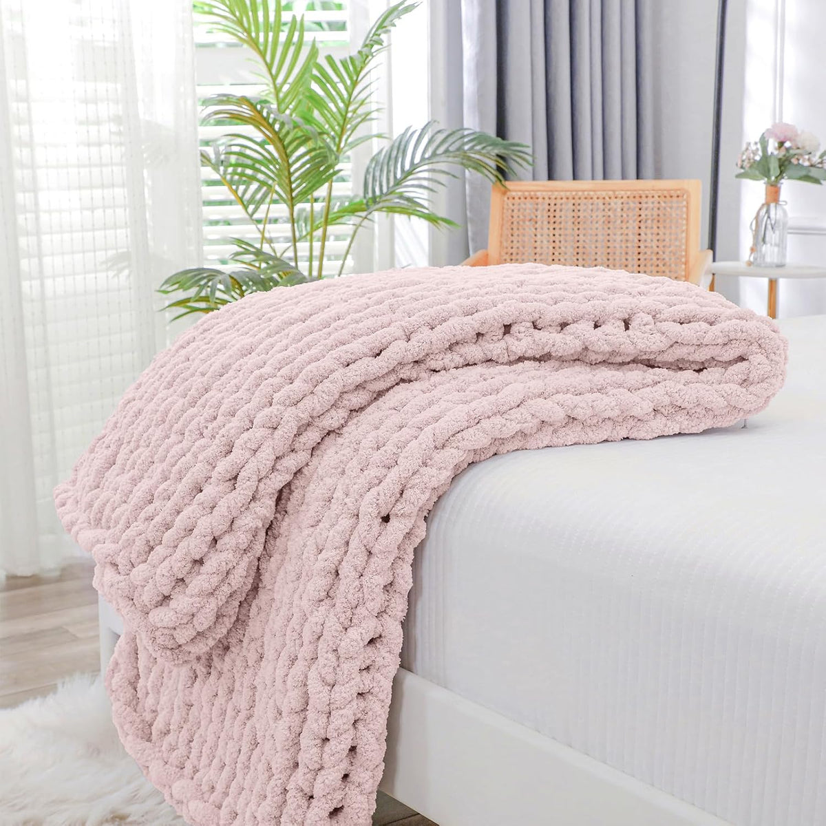 Thick pale pink chunky knit throw folded and draped over a white bed corner with plants behind.
