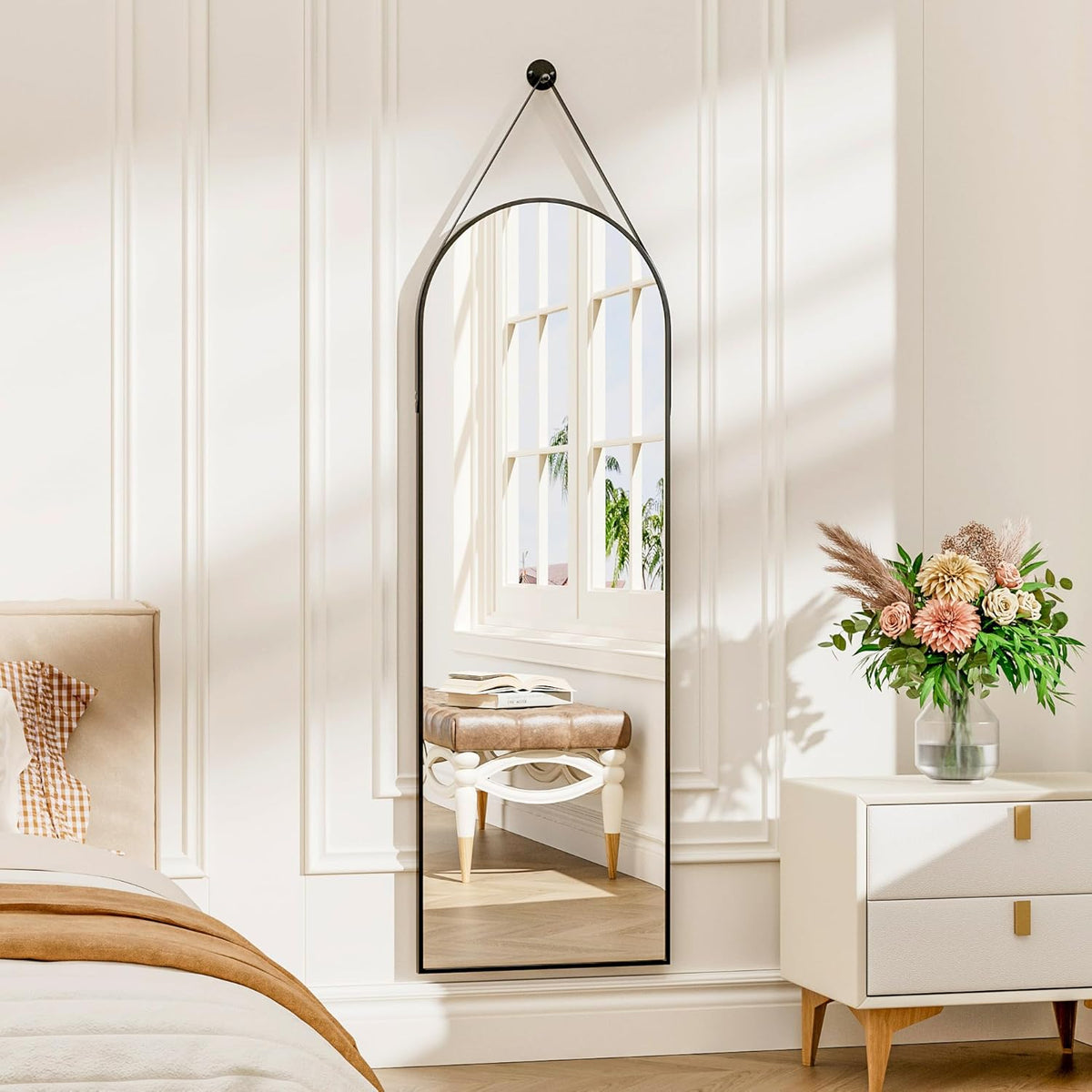 Front view of a tall arched mirror with a thin dark frame mounted on paneled wall, reflecting a window and a tufted stool