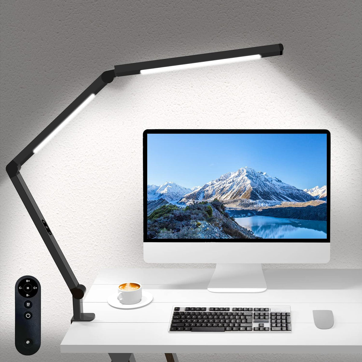 Black dual-arm clamp lamp positioned over a monitor and keyboard on a white desk, remote shown at left.
