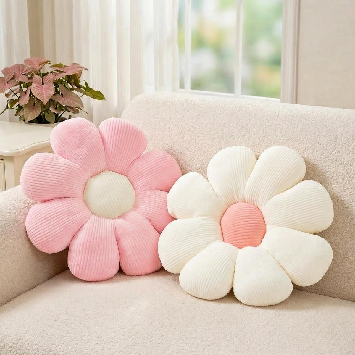 Living-room view of a pink and cream daisy pillow pair propped on a textured beige sofa with a potted plant nearby.