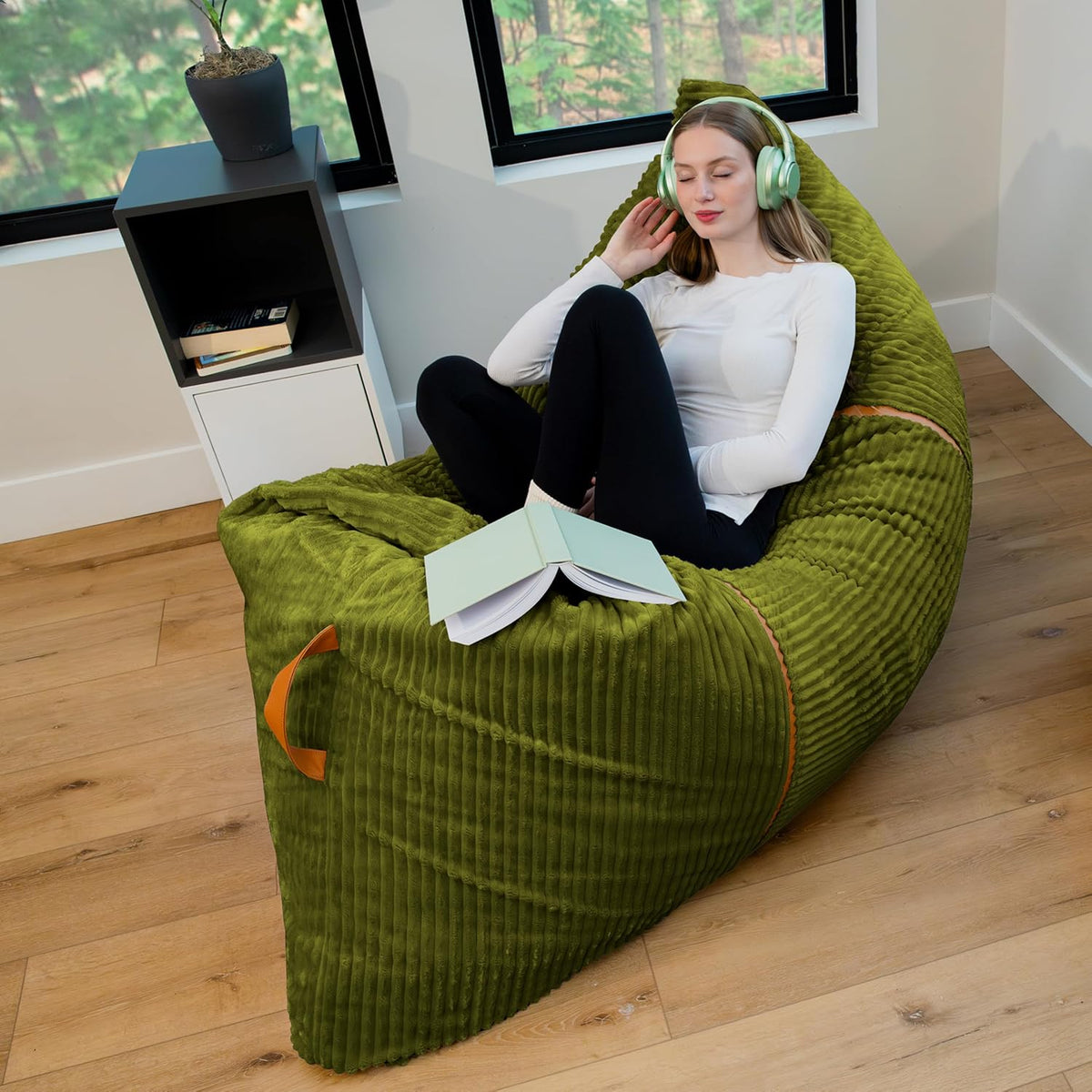 Angled corner view of a moss-green ribbed lounger with orange piping and a woman listening with headphones, book open on the