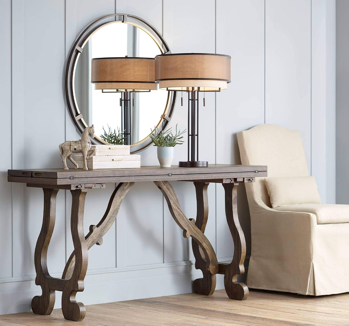 Mirror above a console table reflecting a double-shaded lamp, with a small potted plant, boxes, and wooden horse decor.