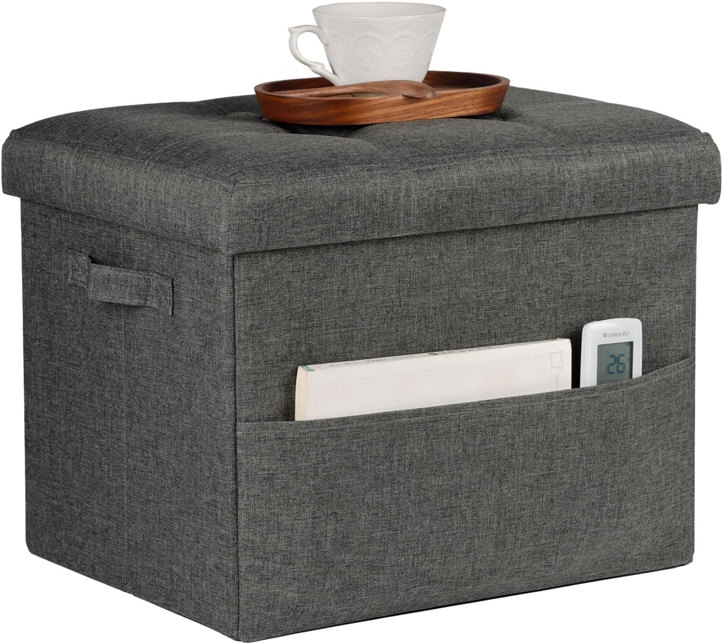 Gray fabric storage ottoman with tufted lid, side handle and front pocket holding a book and remote; wooden tray and teacup