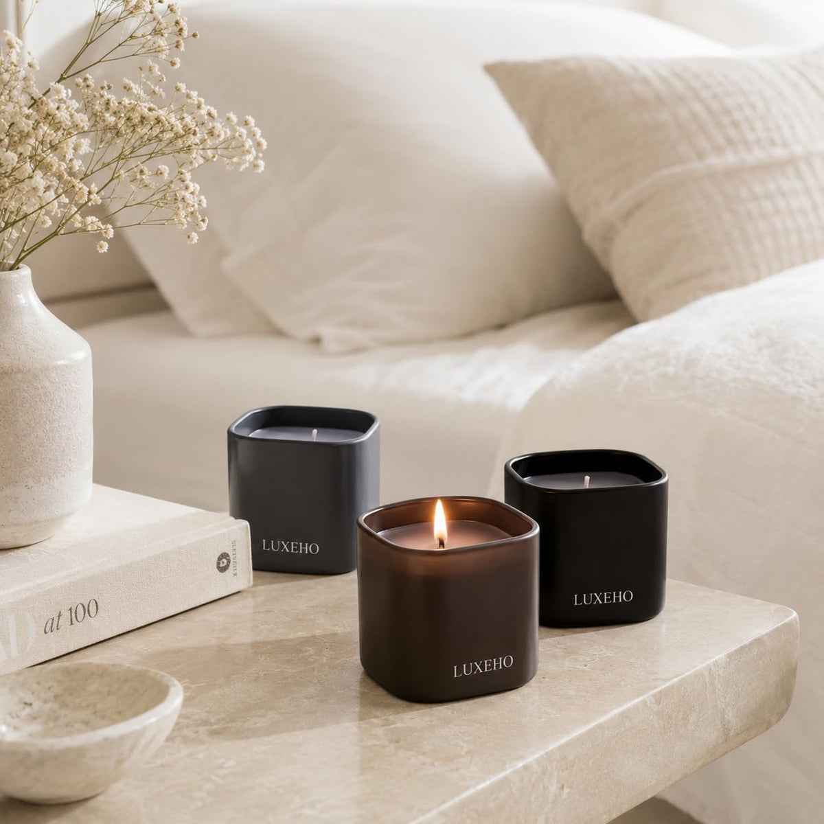 Three square matte-jar candles displayed on a bedside or side table with soft bedding and decor in the background
