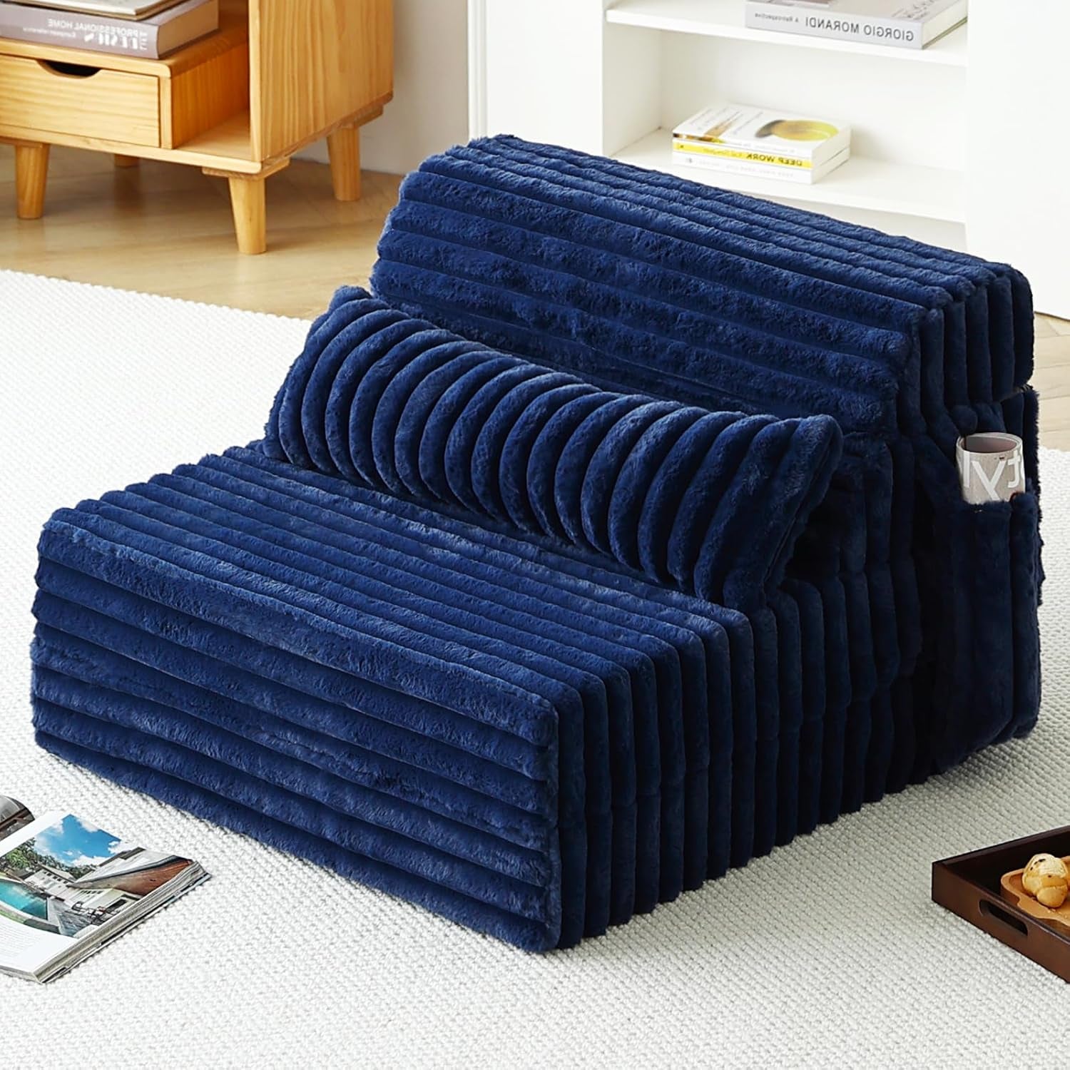Plush navy ribbed folding sofa bed set up as a low chair with bolster pillow and side pocket in a bright room.