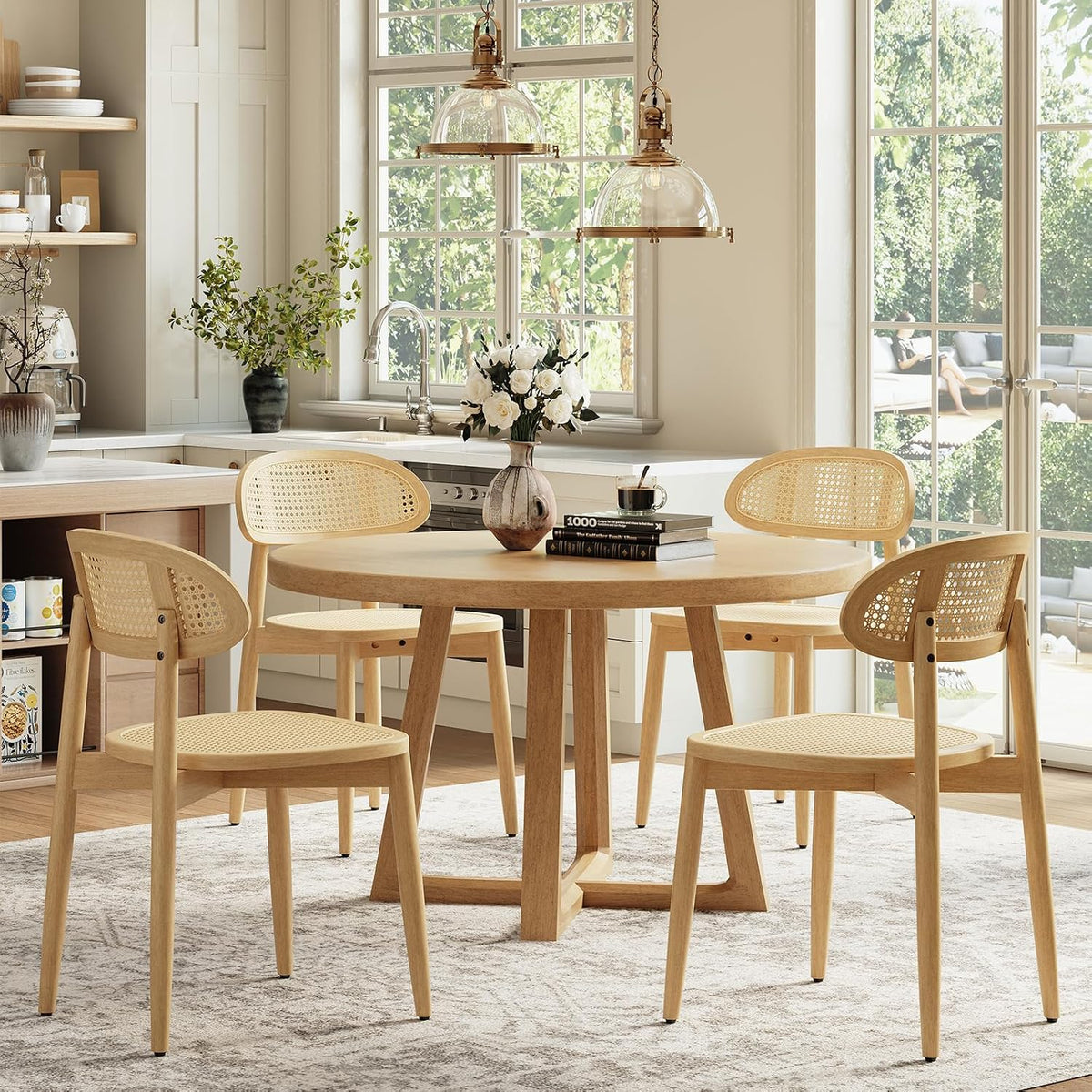 Four light-wood rattan-back dining chairs around a round table near large windows and a kitchen.