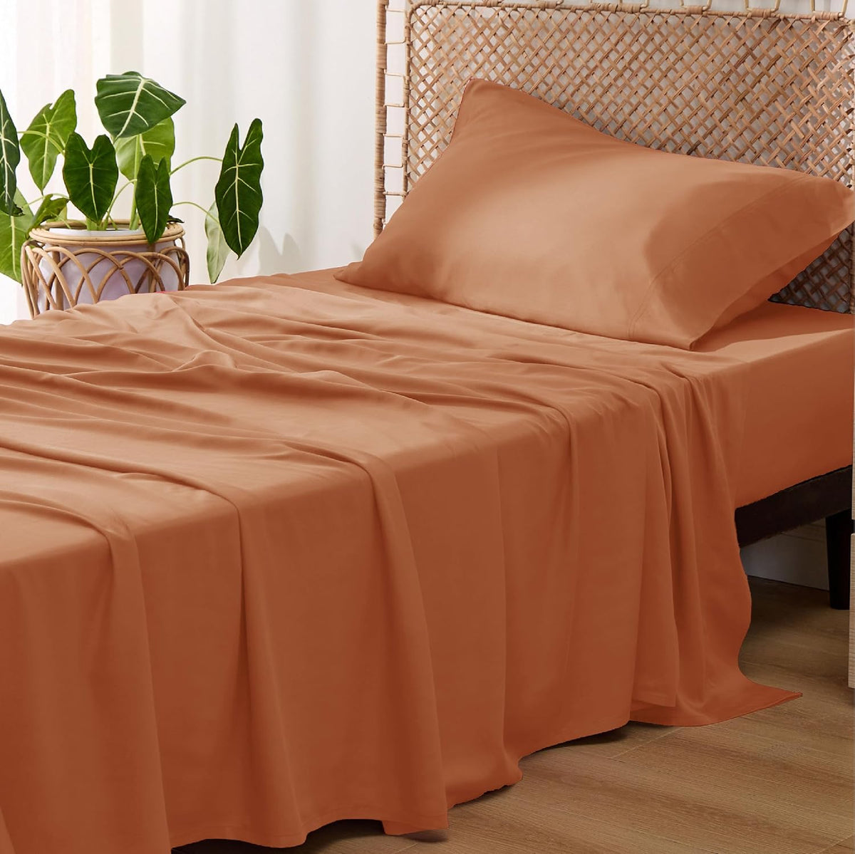Side view of a single bed with warm terracotta sheets and pillow draping to the floor next to a woven headboard and plant.