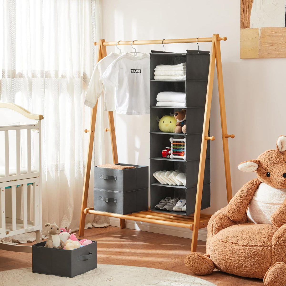 Grey hanging organizer suspended from a wooden garment rack in a nursery setting with folded clothes and toys