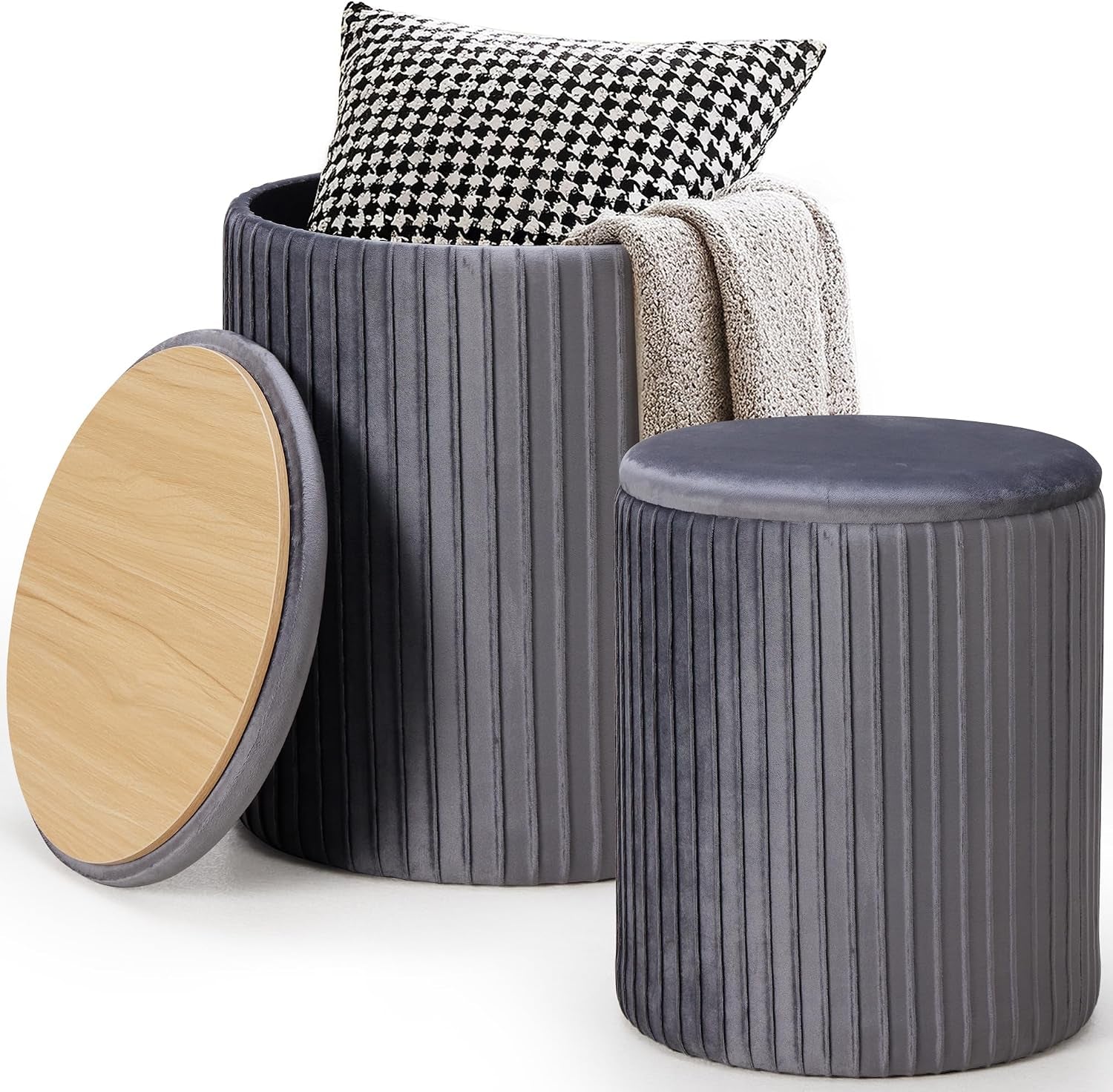 Pair of round gray velvet ottomans with wood lid propped against the larger storage ottoman containing a pillow and throw.