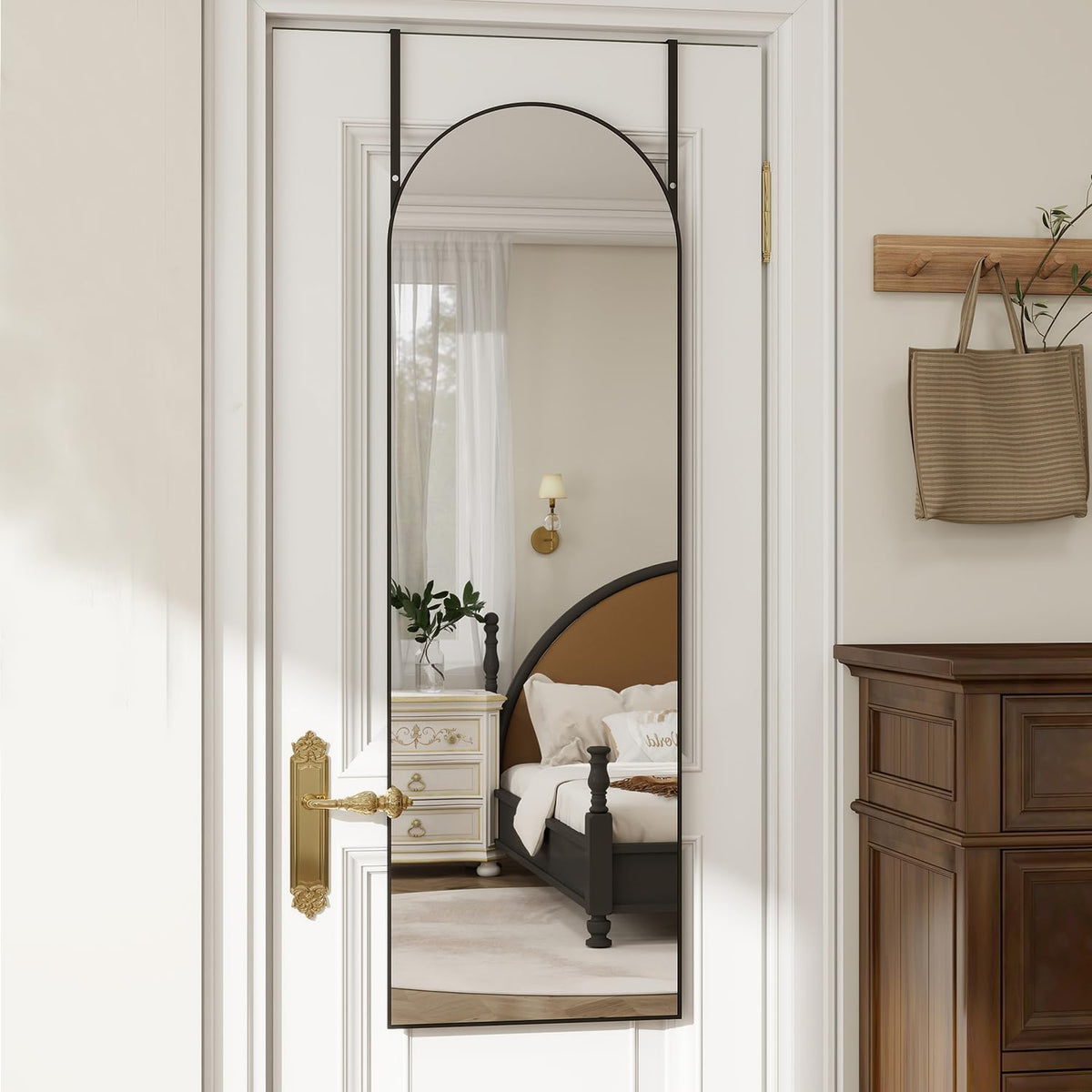 Tall arched mirror hung on a white paneled door showing a bedroom reflection, slim dark frame visible.