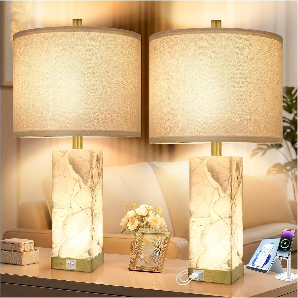 Pair of lit alabaster lamps with square bases, one base showing a USB charging cable plugged in and devices staged nearby.