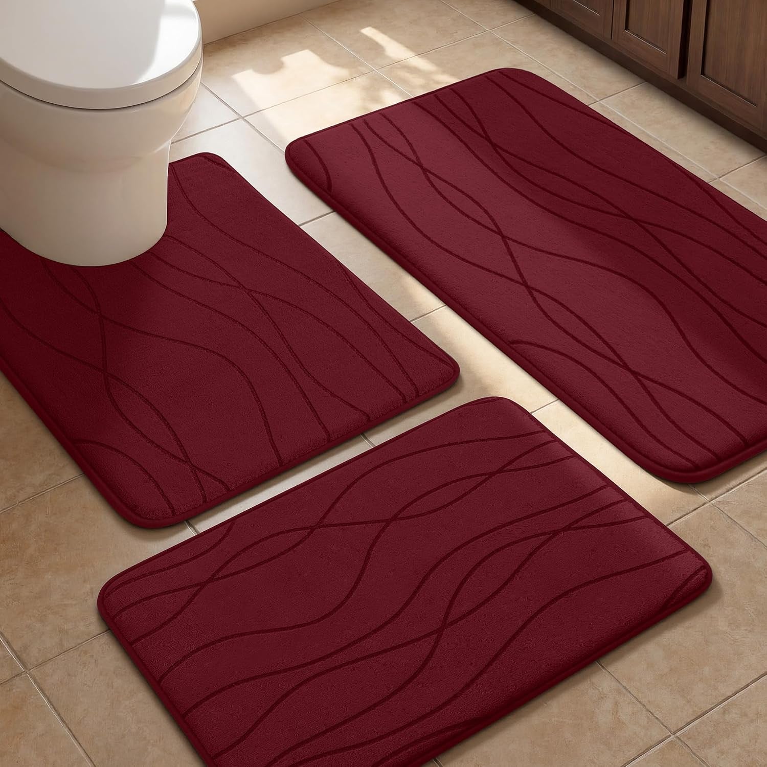 Three burgundy bath mats placed near toilet and tub on tan tile floor