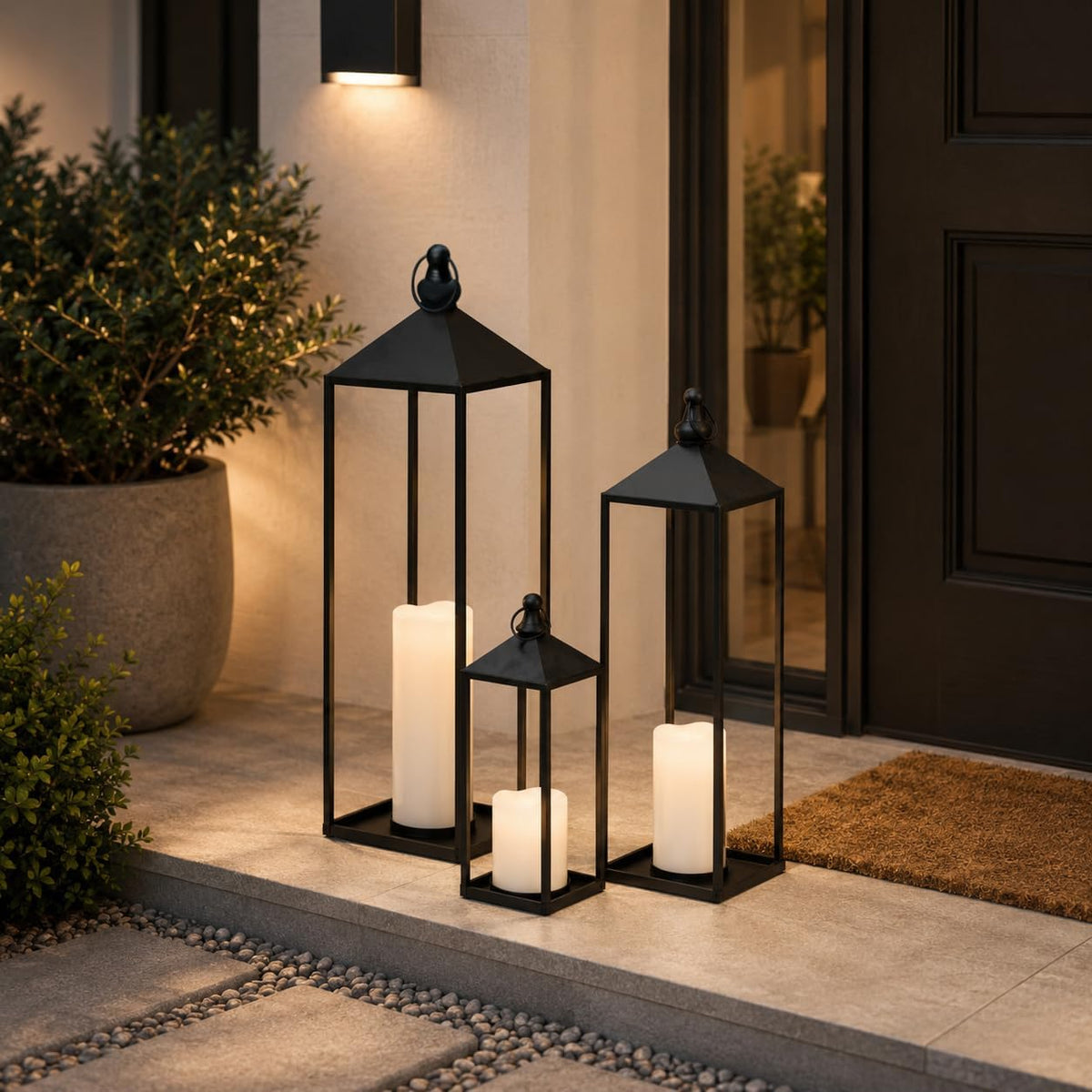 Three lanterns with glowing LED candles grouped on a front porch step beside a doormat at dusk.