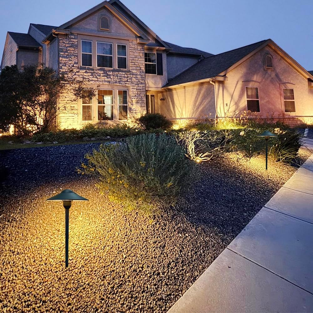 Single black pathway light glowing over gravel and garden beds in front of a lit house at night