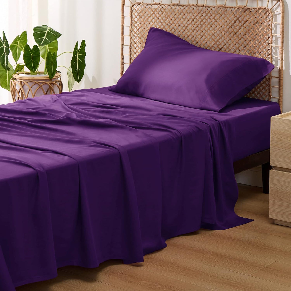 Three-quarter view of a bed dressed in deep purple sheets and a matching pillow against a woven headboard.