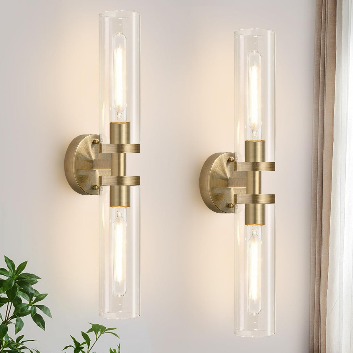 Two vertical brushed-gold wall sconces with clear glass tubes mounted on a light wall beside a curtain.
