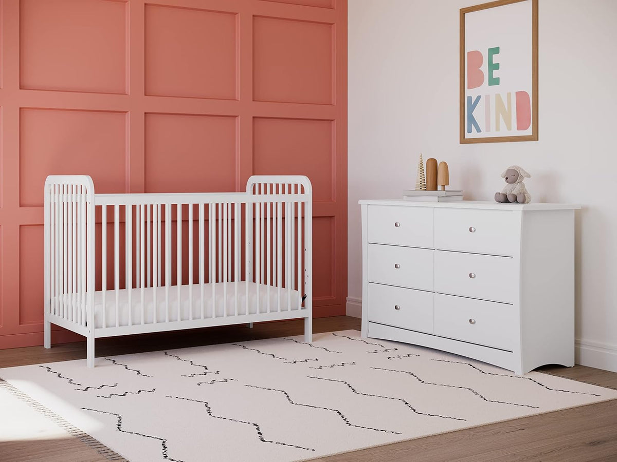 White dresser beside a white crib in a nursery with a coral accent wall, angled lifestyle view