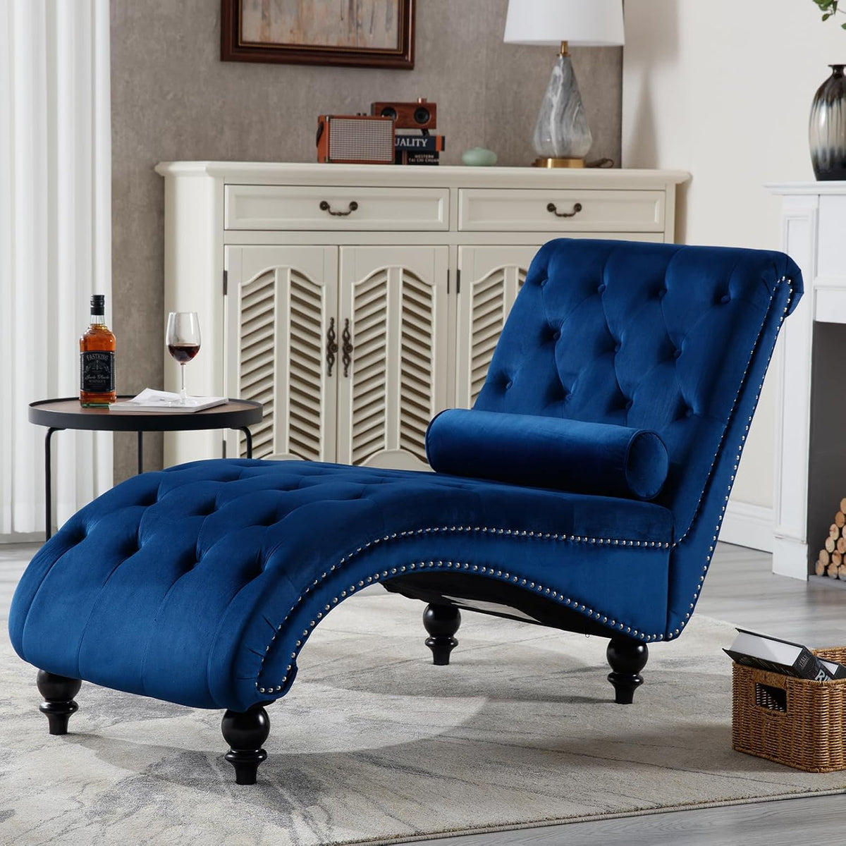 Blue tufted S-shape chaise angled left with bolster pillow, nailhead trim, turned black legs in a styled living area.