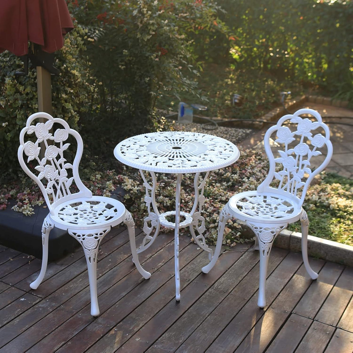 White cast-aluminum bistro table with umbrella hole and two matching floral-back chairs on a wooden deck.