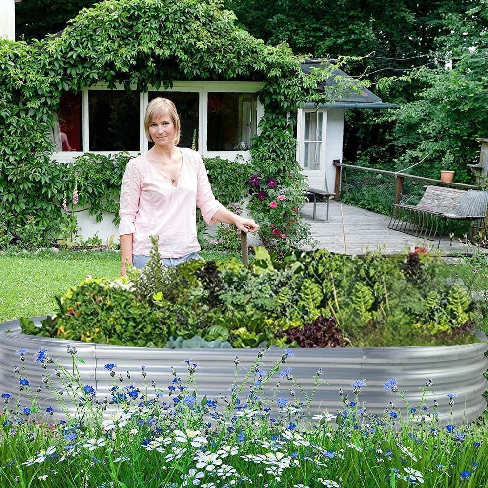 Woman standing behind a large oval galvanized raised garden bed filled with plants in a backyard setting.