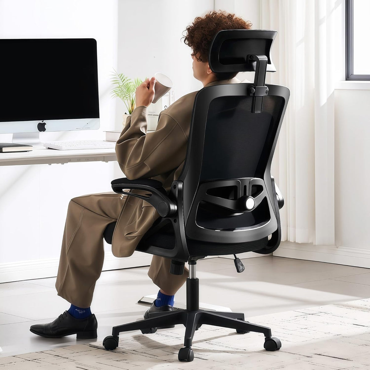 Black mesh office chair rear three-quarter view with person seated at a white desk in a bright office