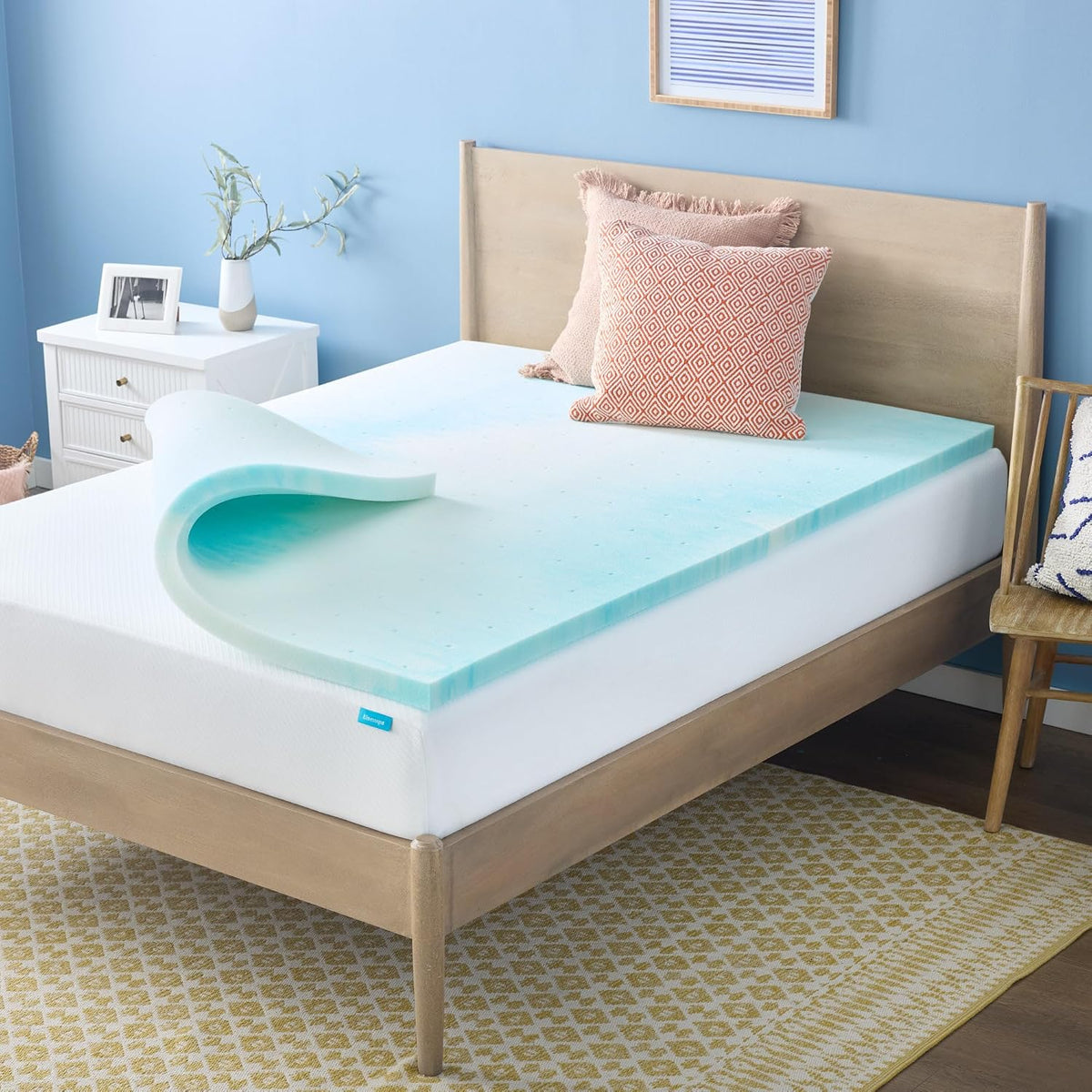 Side-angle view of a light blue gel foam topper laid on a wooden bed frame with patterned rug and pillows.