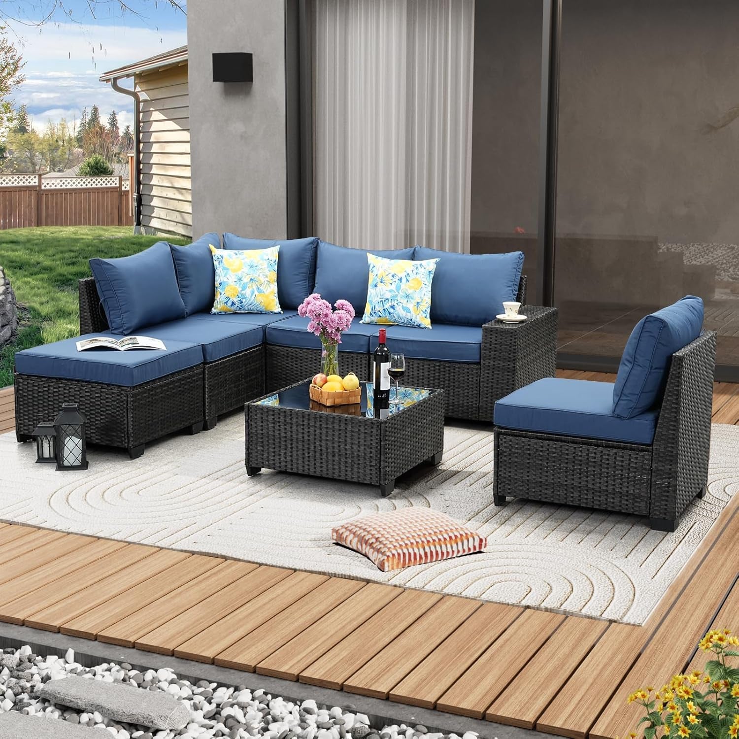 Rattan patio sectional arranged on a deck with blue cushions, decorative pillows and a square glass coffee table.