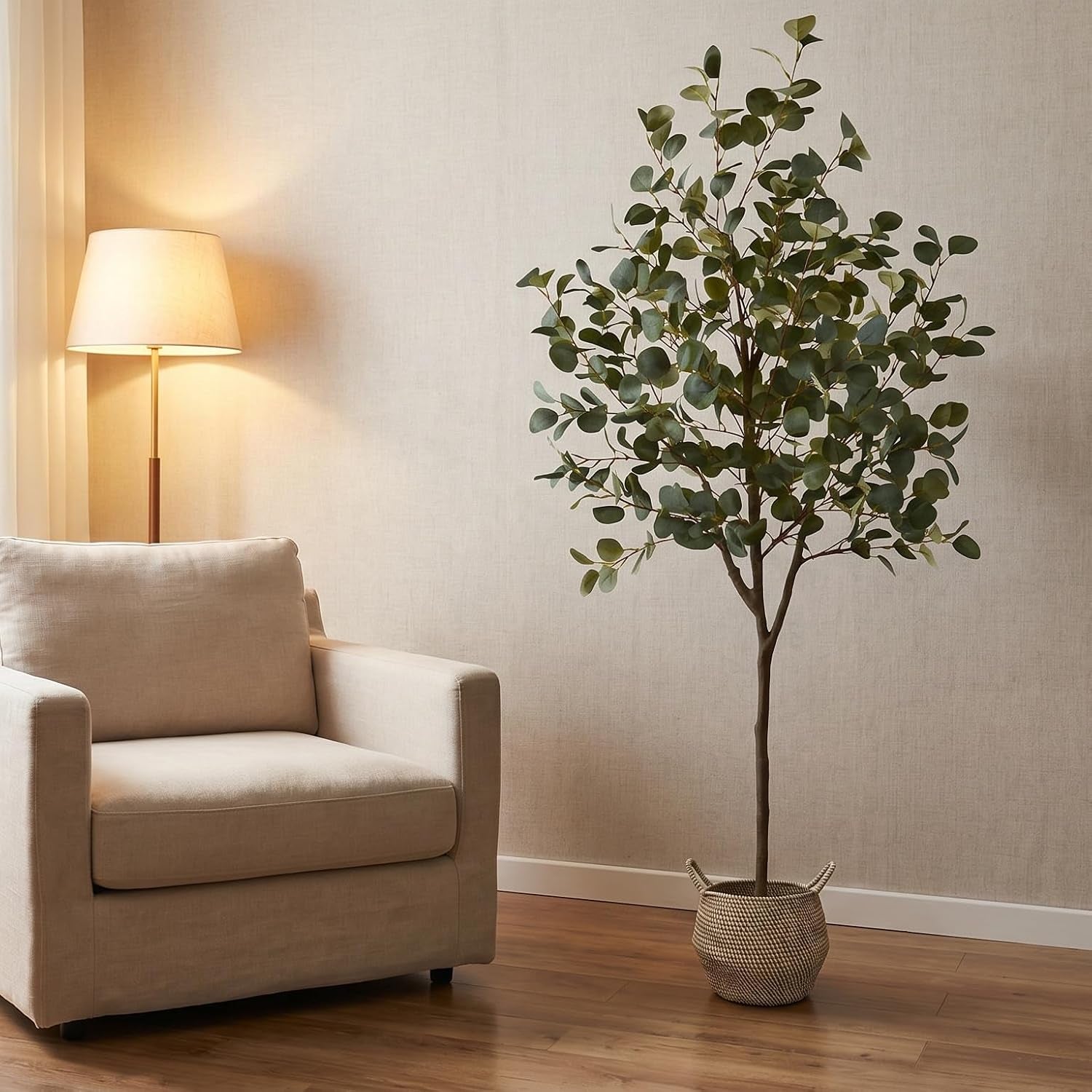 Eucalyptus tree in a textured basket beside a beige armchair and lit floor lamp on wood floor.