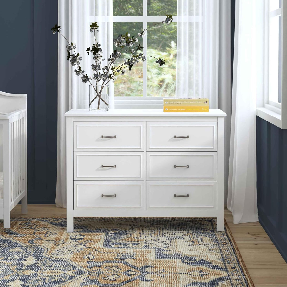 White six-drawer dresser under a window with vase and books on top in a nursery