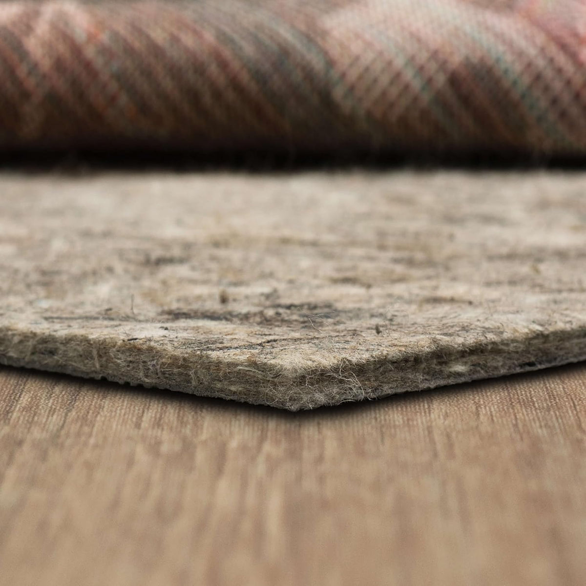 Low-angle close view of a felt rug pad corner resting on a wood floor, showing compressed edge and loose fibers.