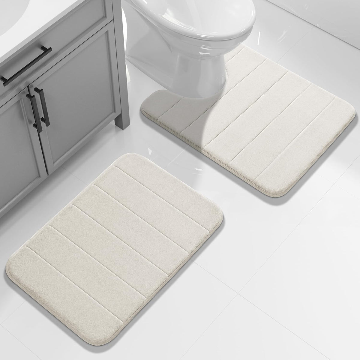 Light beige bathroom mats on a glossy tiled floor, one U-shaped around the toilet and one rectangular by the vanity.