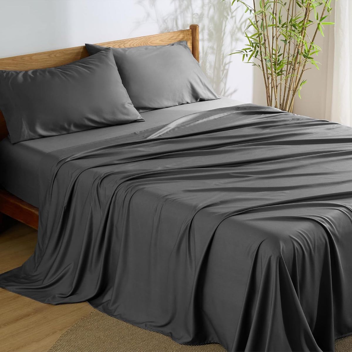 Dark gray sheets and two matching pillowcases draped over a wooden bed with a bamboo plant at the head.