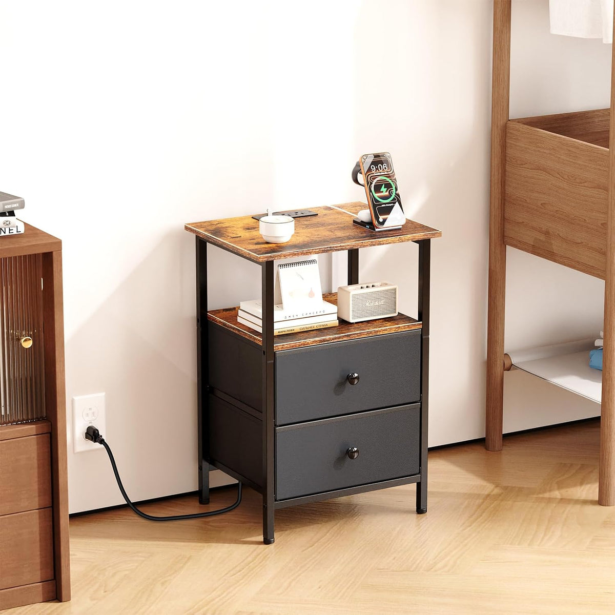 Nightstand beside a bed with a phone charging on top and its cord plugged into a wall outlet.
