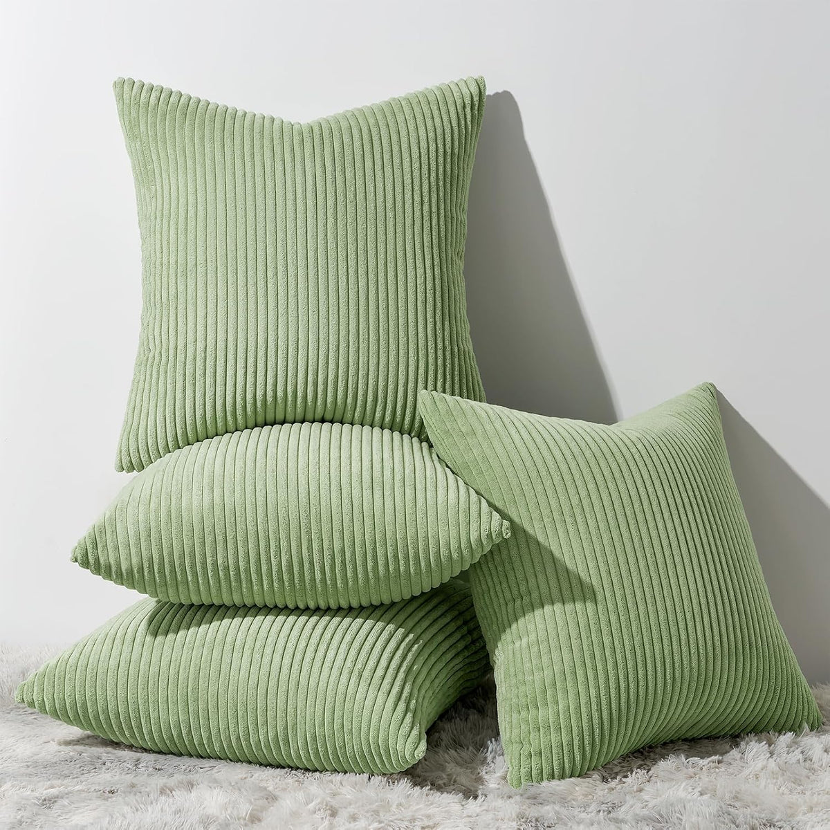 Stack of four sage green ribbed corduroy pillows on a plush rug against a white wall