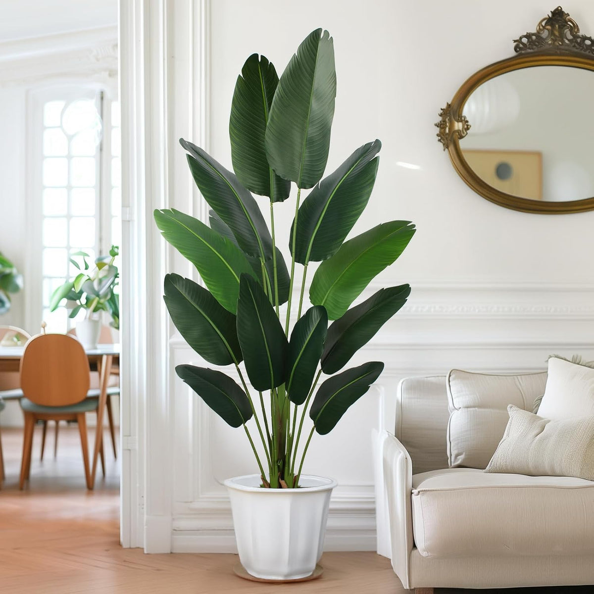 Single artificial bird-of-paradise in a white fluted planter next to a beige sofa and gold oval mirror