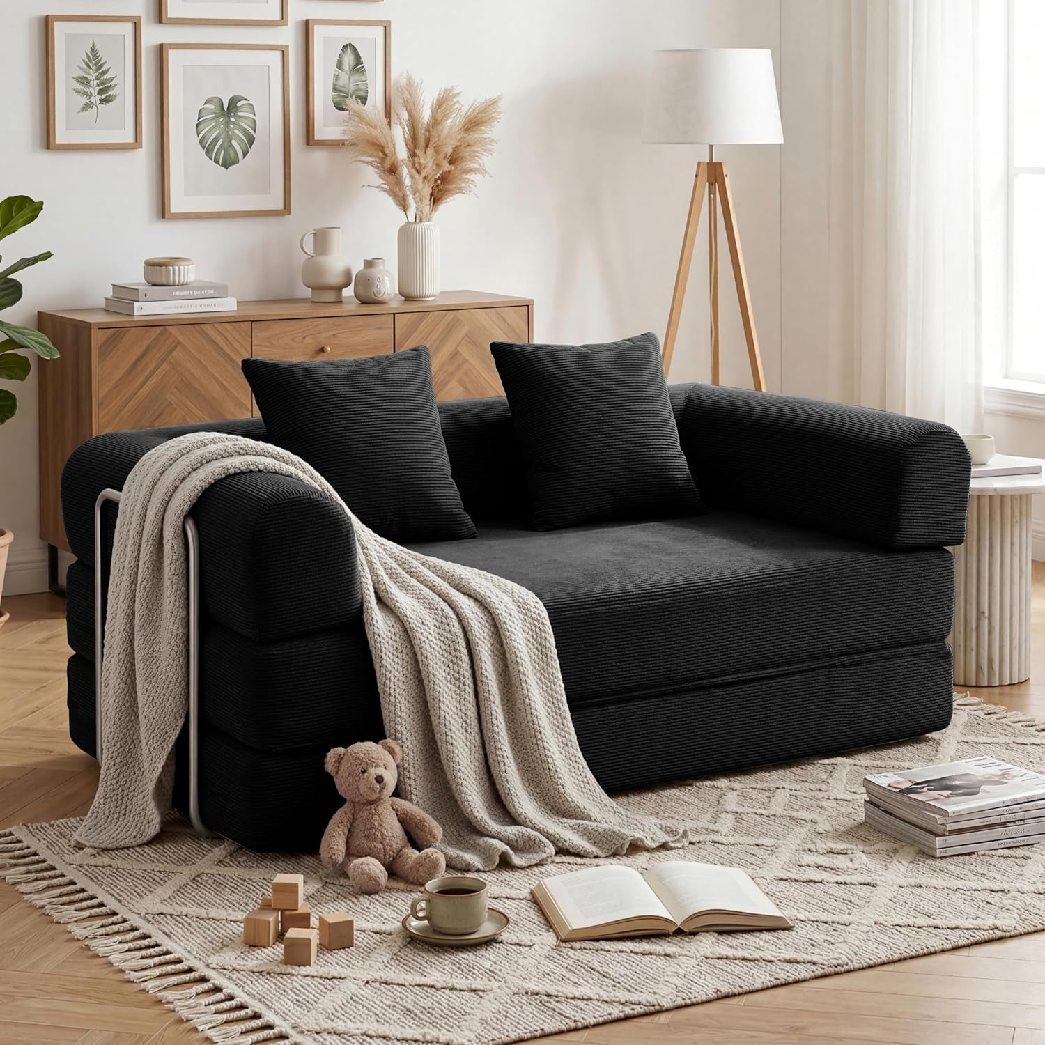 Black corduroy sofa with pillows, a knit throw over one arm, and a teddy on the rug beside an open book