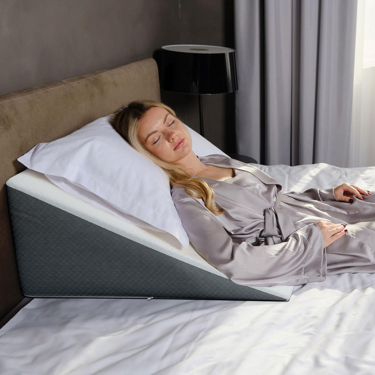 Woman sleeping on a gray-and-white wedge pillow on a bed with a black lamp and gray curtains behind.