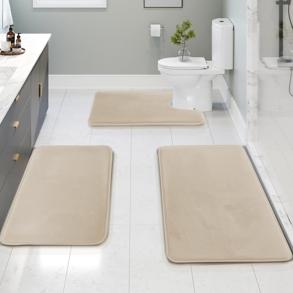 Bathroom scene with three beige memory-foam mats placed by a vanity, shower and around the toilet on white tile.