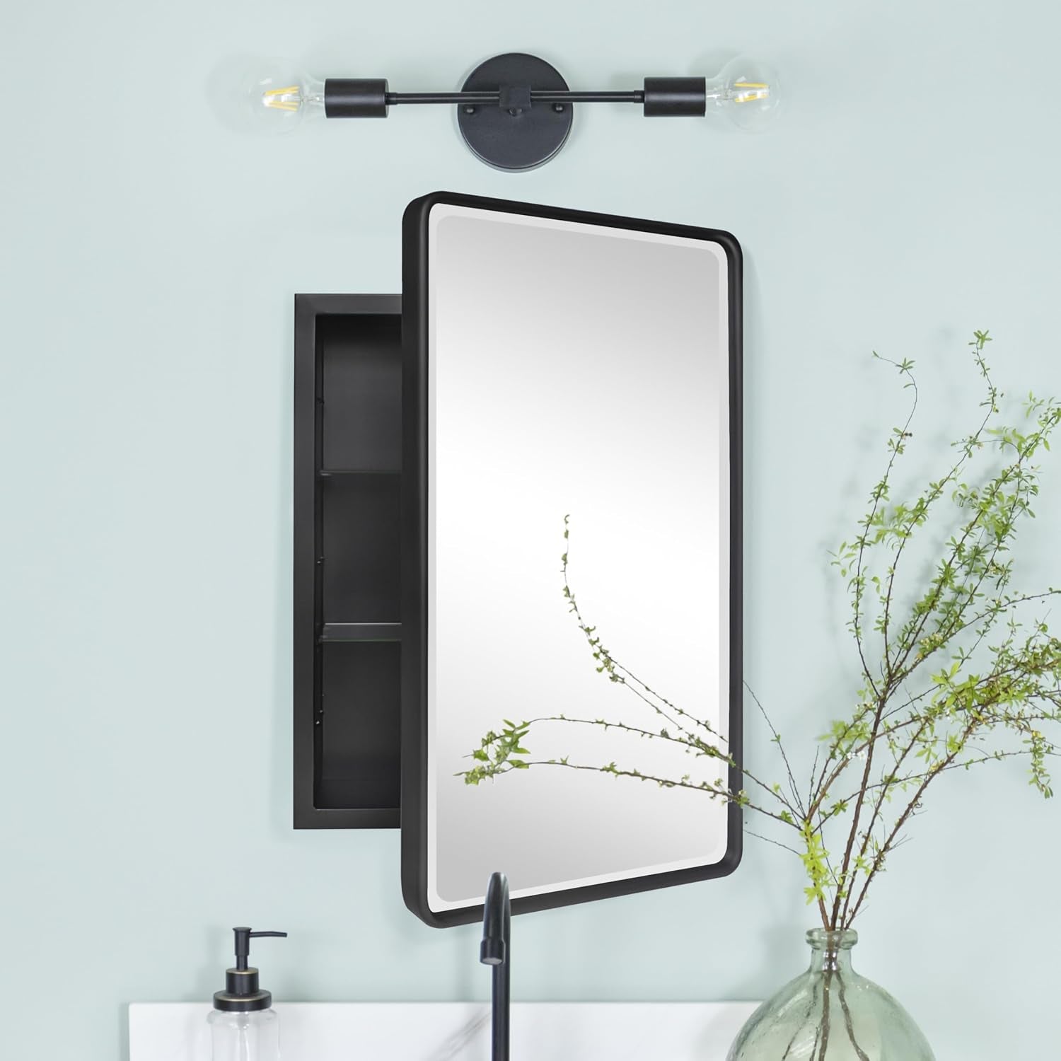 Cabinet mounted on a pale blue wall beside a black faucet and a vase of leafy branches, with a two-bulb sconce above.