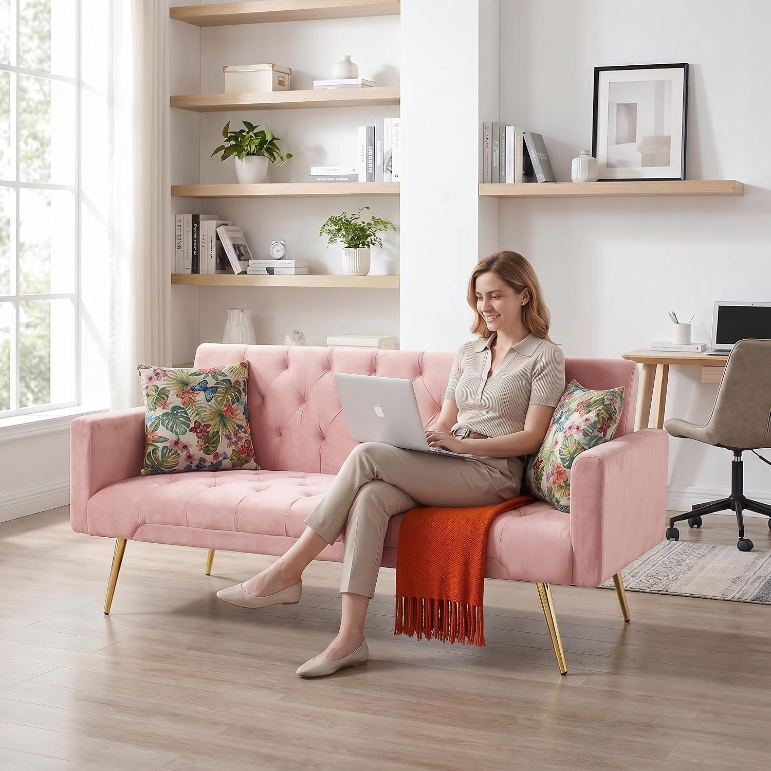 Woman sitting on a pink velvet sofa using a laptop, with floral pillows and an orange throw draped over the seat.