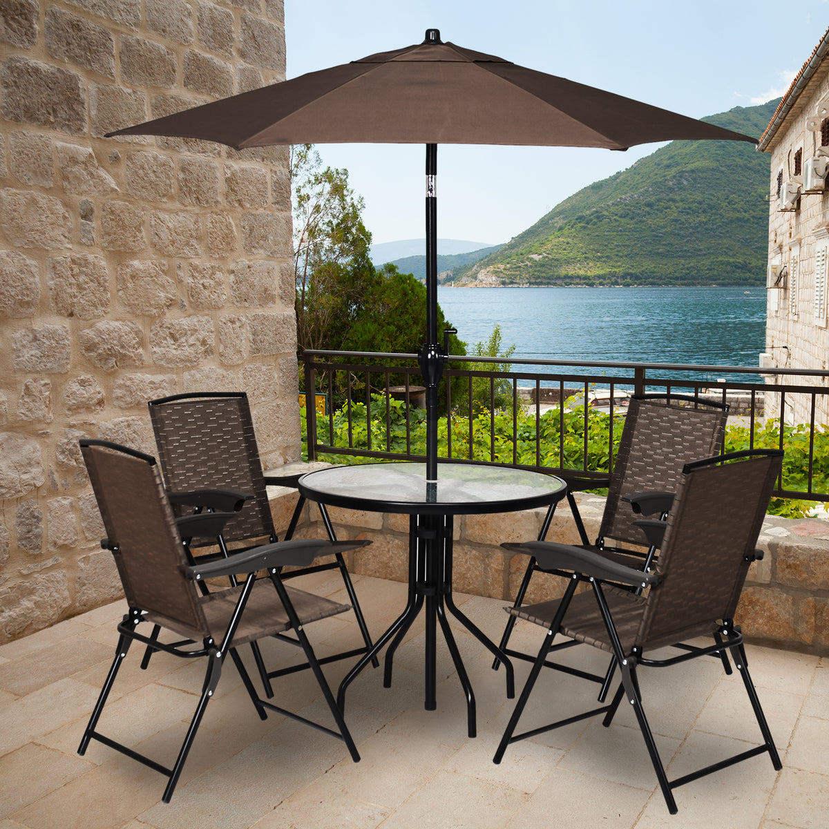 Four folding chairs surround a round table beneath a patio umbrella on a stone terrace overlooking the water.