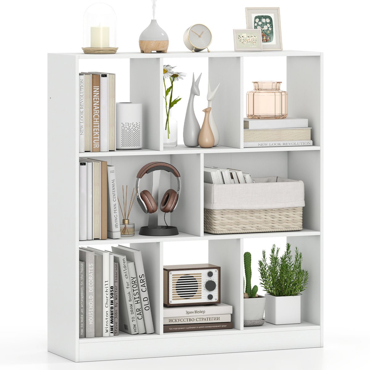 White open bookcase filled with books, headphones, baskets, plants, and decorative objects against a white backdrop.